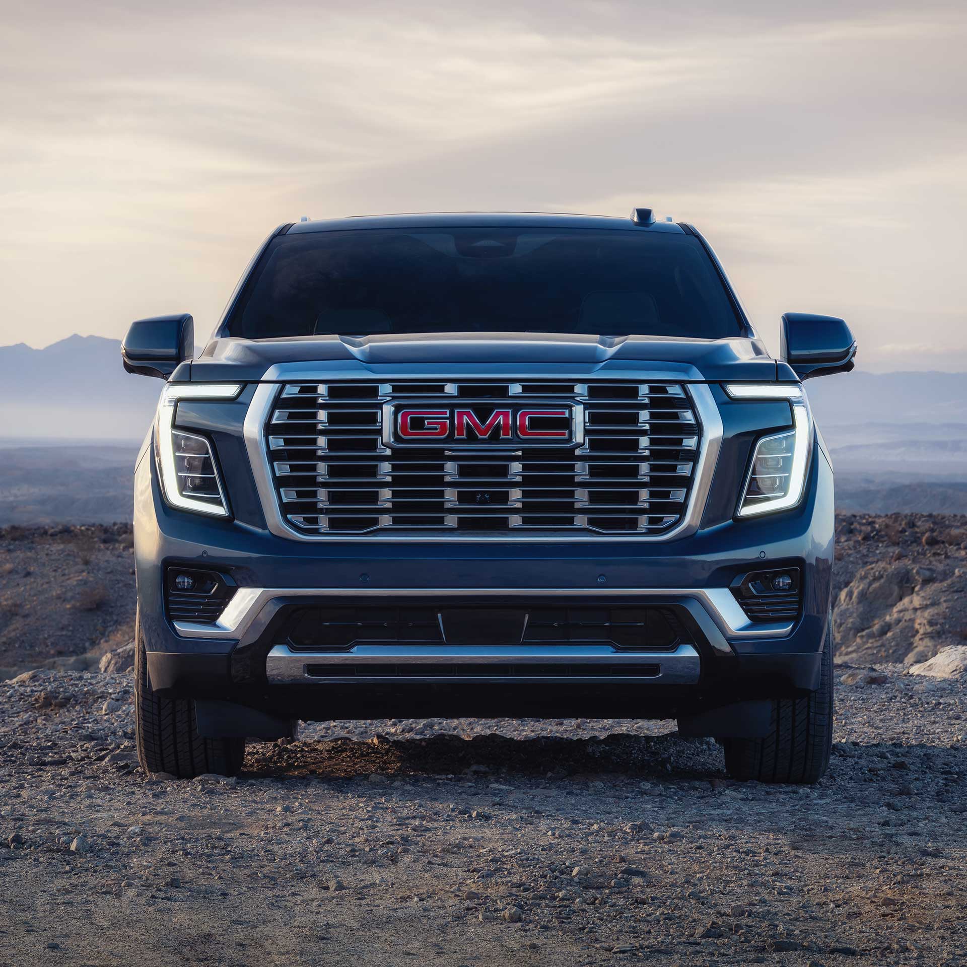 The Front View of a Dark Blue GMC Yukon SUV with a Prominent Grille and GMC Logo, Parked On a Dirt Path At Sunset