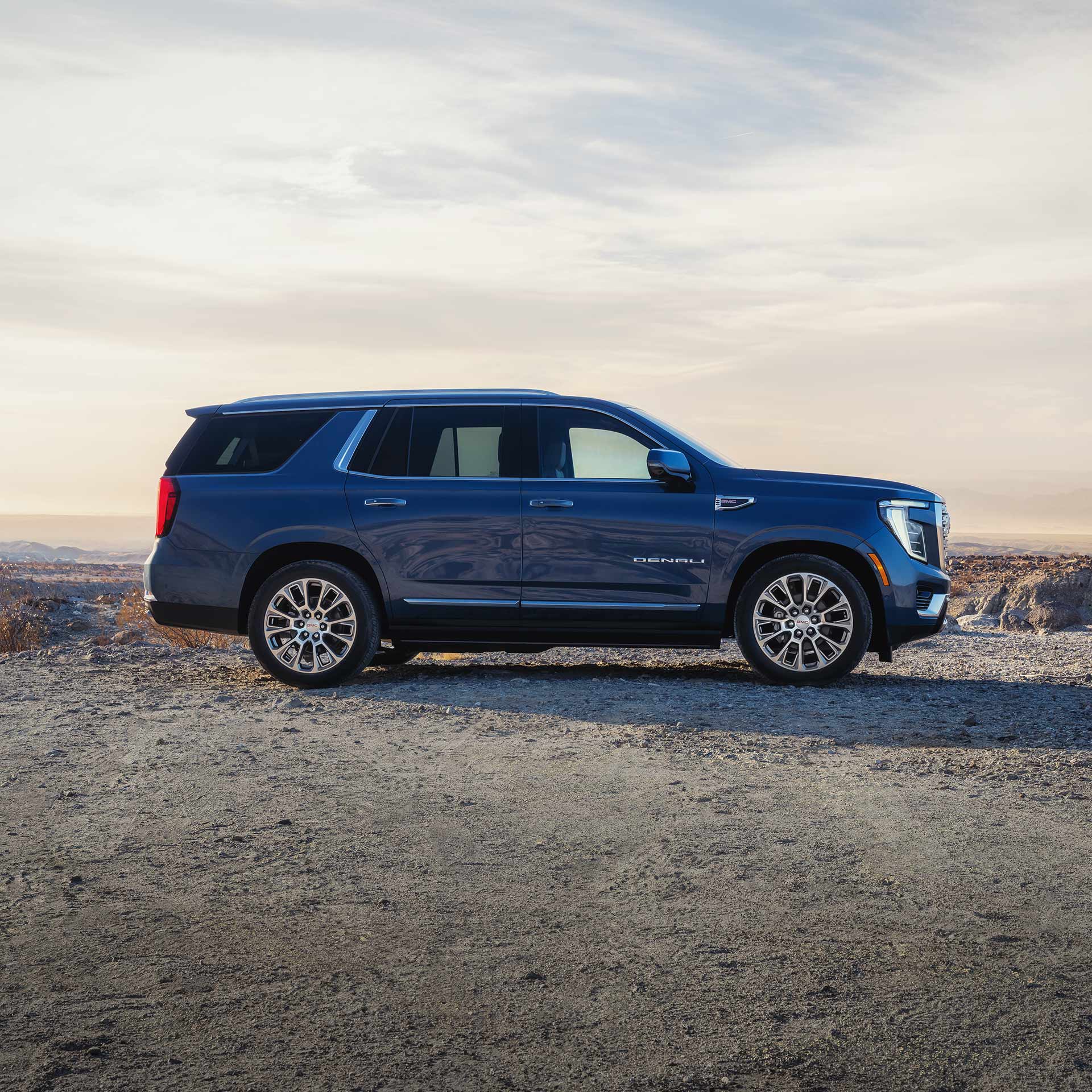 A Dark Blue GMC Yukon Denali SUV Is Parked On a Dirt Road with Mountains In the Background Under a Cloudy Sky