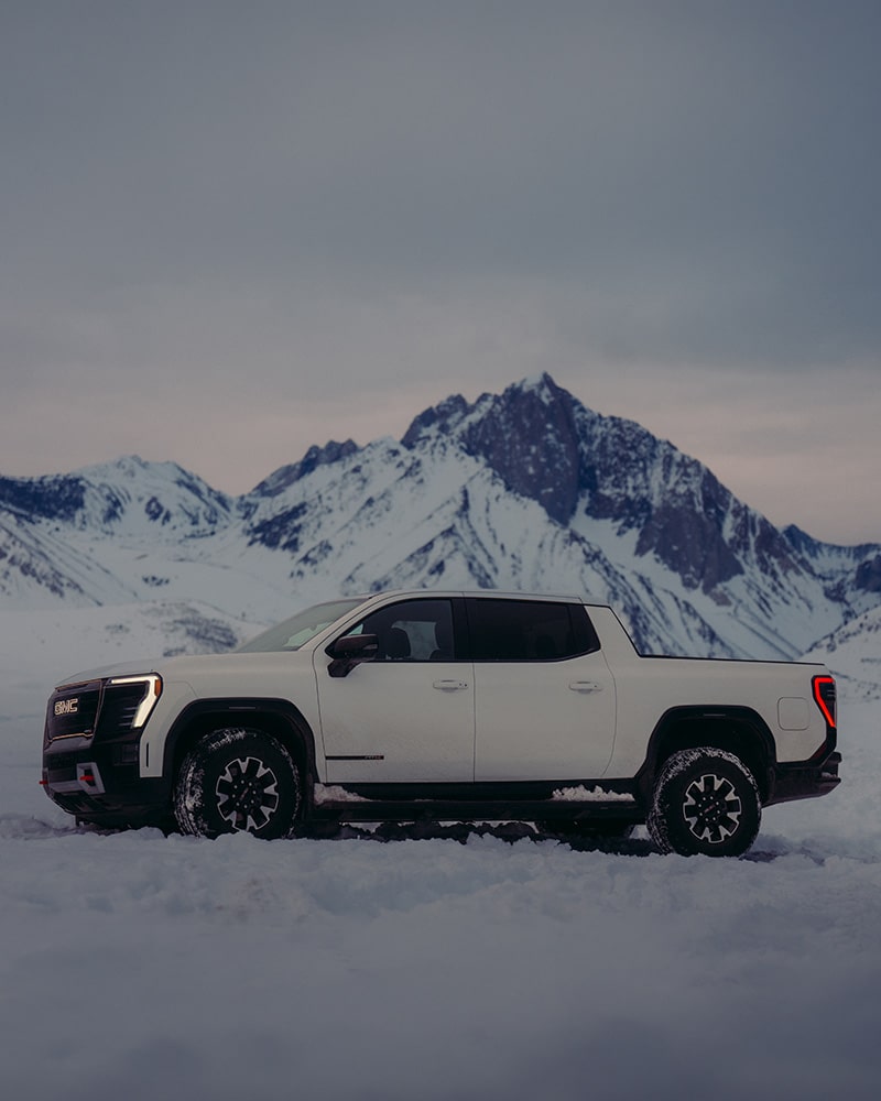 A White GMC Canyon AT4X Truck Is Parked in Deep Snow with Majestic Snow-Capped Mountains in the Background under a Dramatic Sky