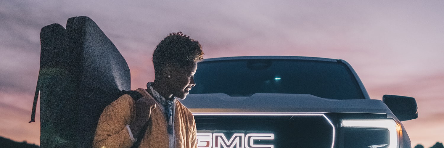 L. Renee Blount Wearing a Gold Jacket and Gray Pants Stands Beside the Illuminated Grille of a GMC Truck Carrying a Dark Bag over Their Shoulder at Dusk