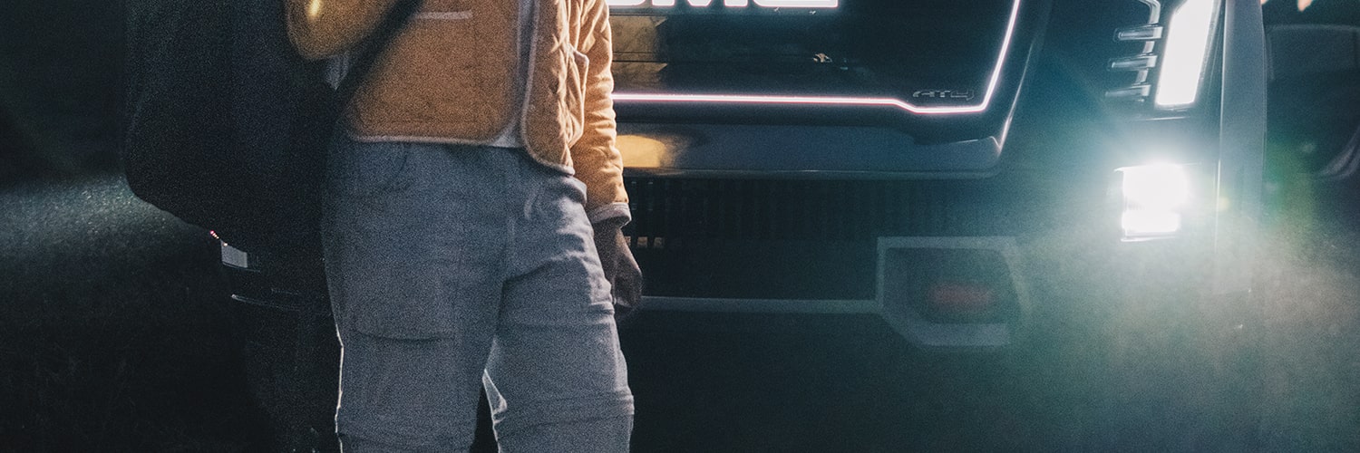L. Renee Blount Wearing a Gold Jacket and Gray Pants Stands Beside the Illuminated Grille of a GMC Truck Carrying a Dark Bag over Their Shoulder at Dusk