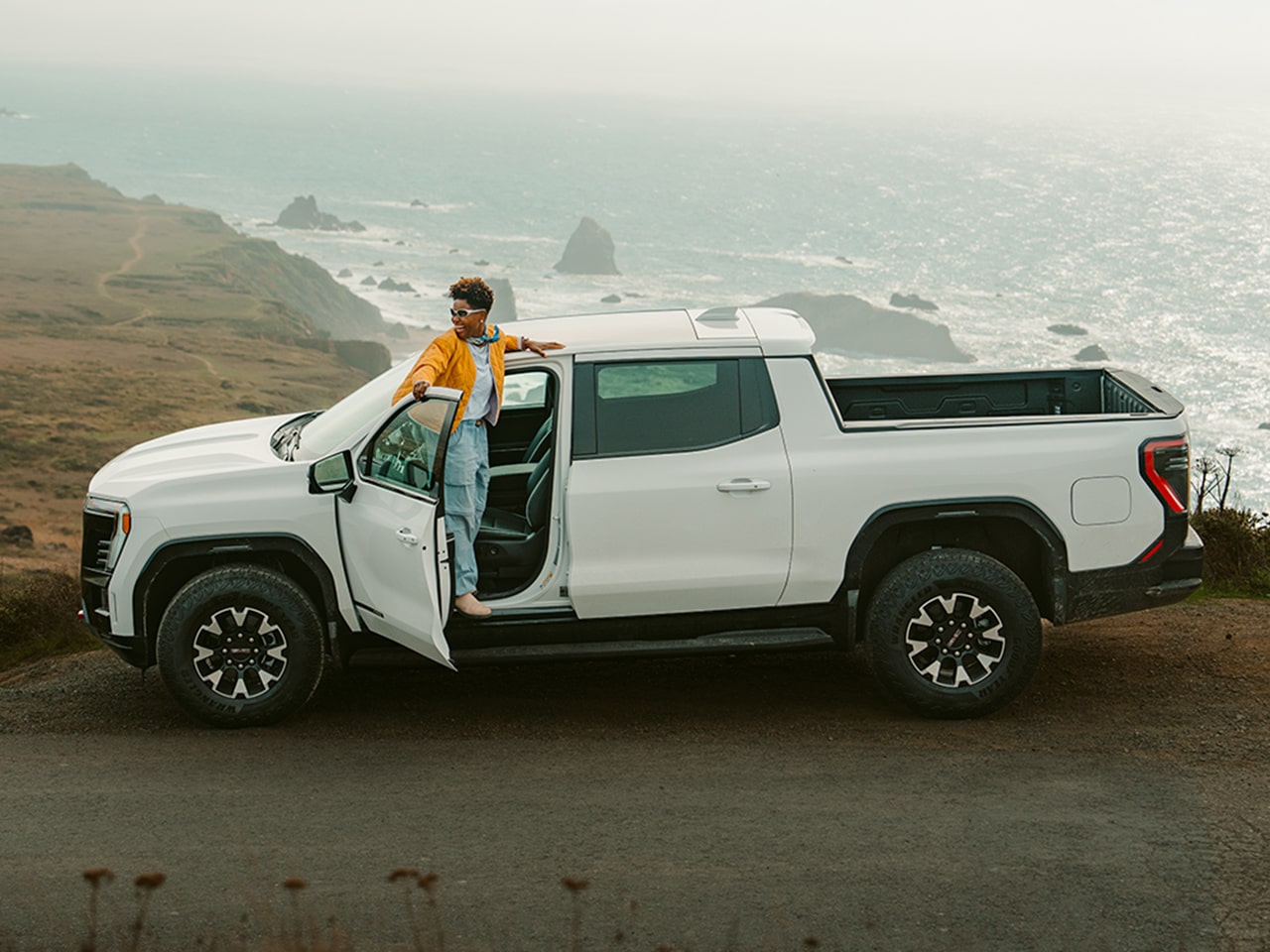 L. Renee Blount in a Yellow Jacket Leaning out of the Open Driver's Door of a White GMC Truck Overlooking the Ocean