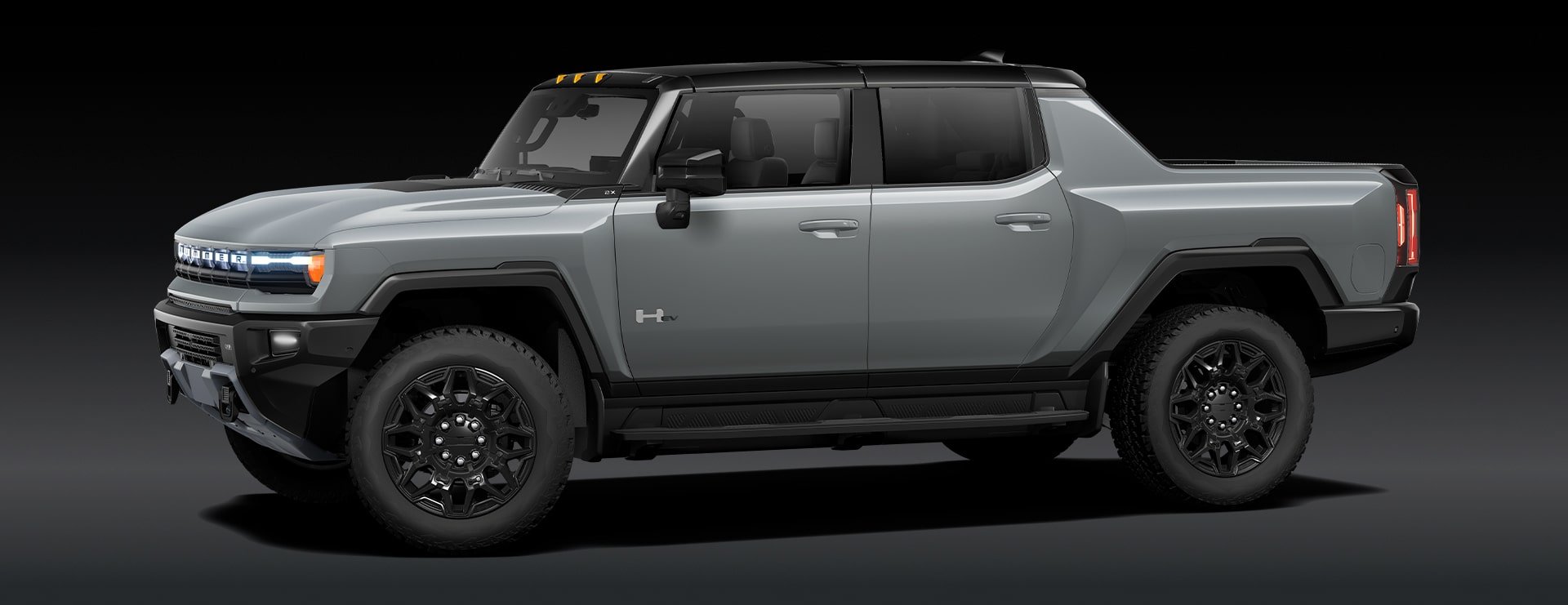 A Gray GMC HUMMER EV Pickup Truck Is Parked From a Front Three-Quarters Angle Against a Dark Background