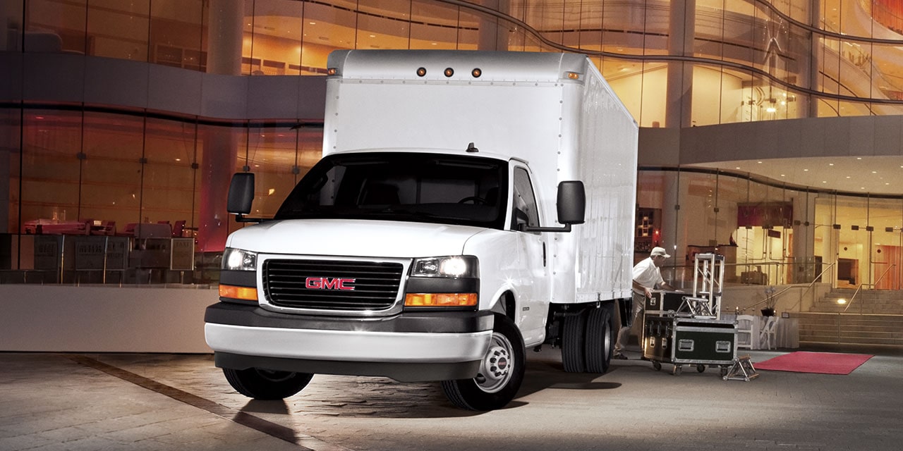 Two GMC Commercial Savana Cutaway Vans Parked in Front of a Large Lite up Building at Night