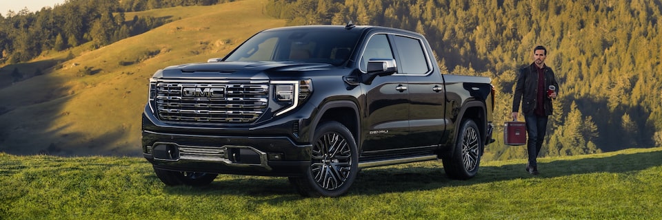 Man Walking Away From Pacific Blue Metallic GMC Sierra Denali Parked Off-Road With Hills and Trees