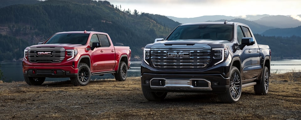 Pacific Blue Metallic and Cardinal Red GMC Sierra Pickup Trucks Parked Off-Road With Lake and Mountains