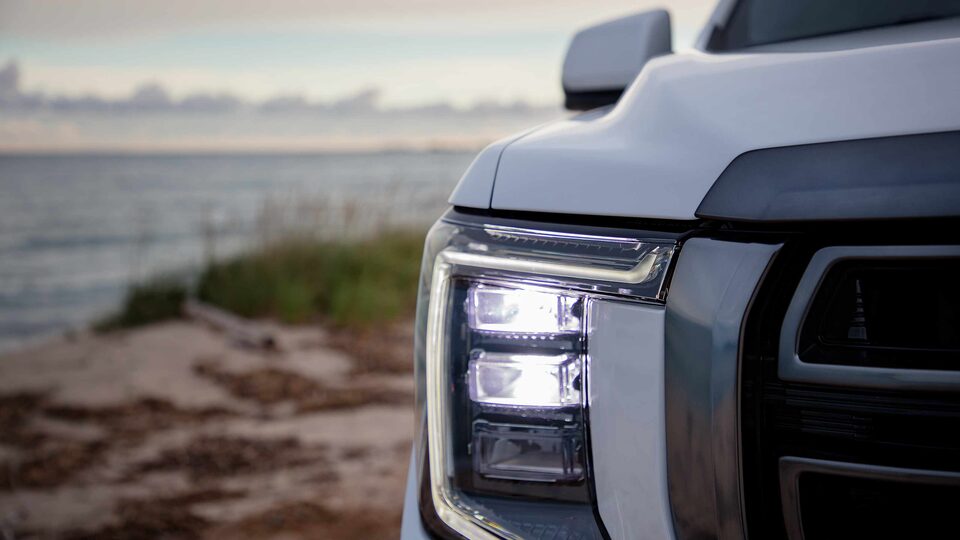 Headlight on the White GMC Yukon AT4 Parked on Beach With Water and Sunset