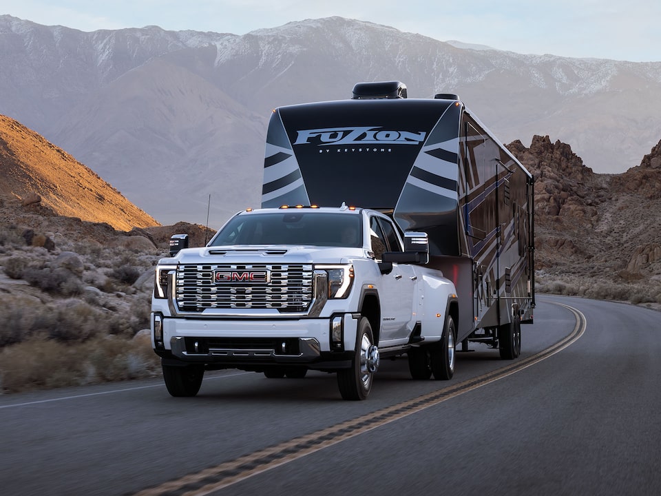Front Three-Quarters View of a 2025 GMC Sierra HD Pickup Truck Hauling a Trailer on an Open Road Surrounded by Mountains