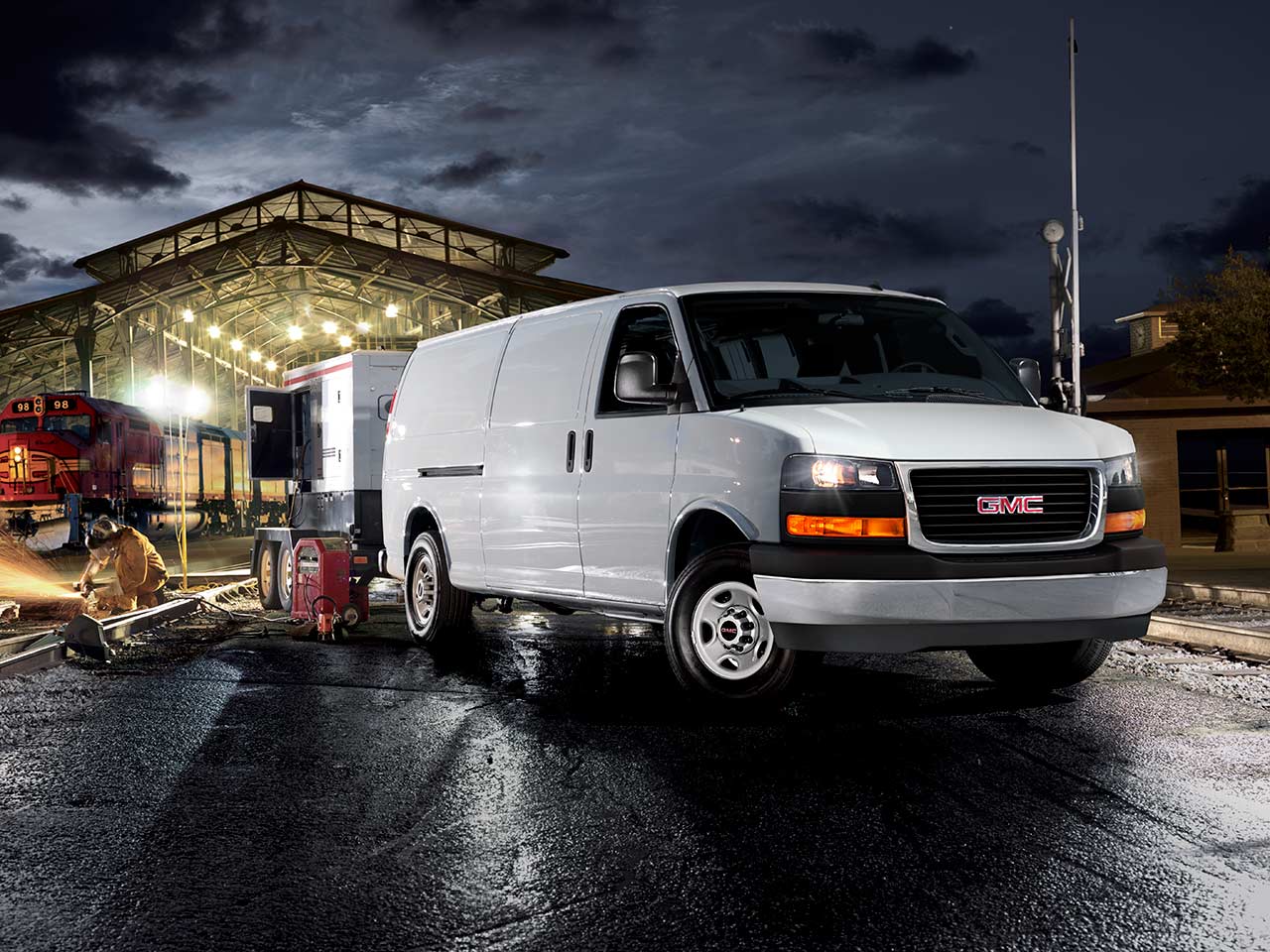A White 2026 GMC Savana Cargo Van with an Attached Generator Trailer on Train Tracks at Night