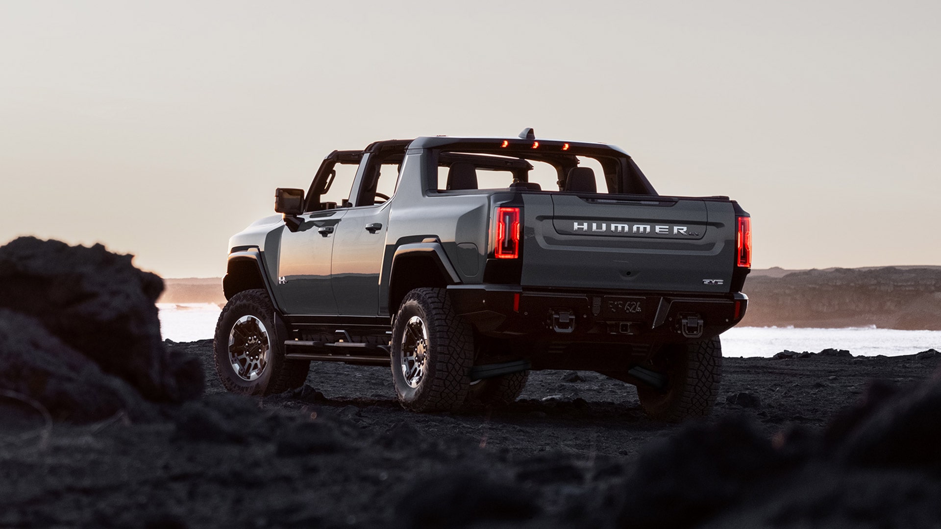 Rear Three-quarter View of the 2026 GMC HUMMER EV Pickup Truck with the Top Off Parked on a Rocky Beach