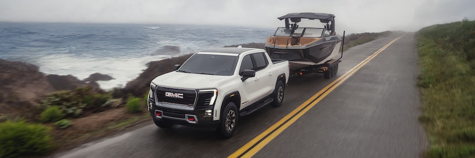 Three-quarters View of a White 2026 GMC Sierra EV Pickup Truck Towing a Boat near an Ocean Road
