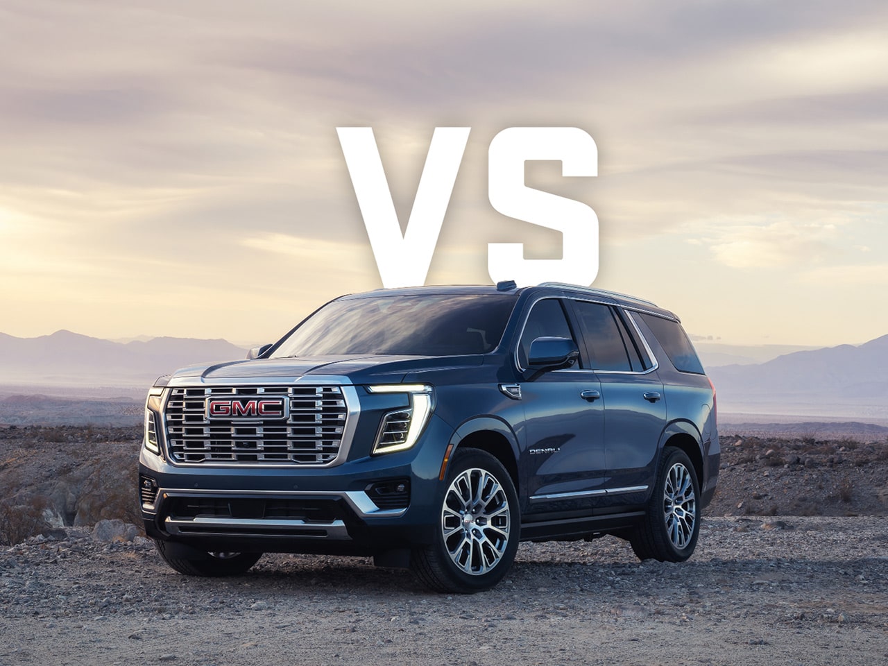 A Dark Blue GMC Yukon Parked in the Desert Surrounded by Mountains with Large Bold Text Overlay Reading 'VS' Centered Above the SUV