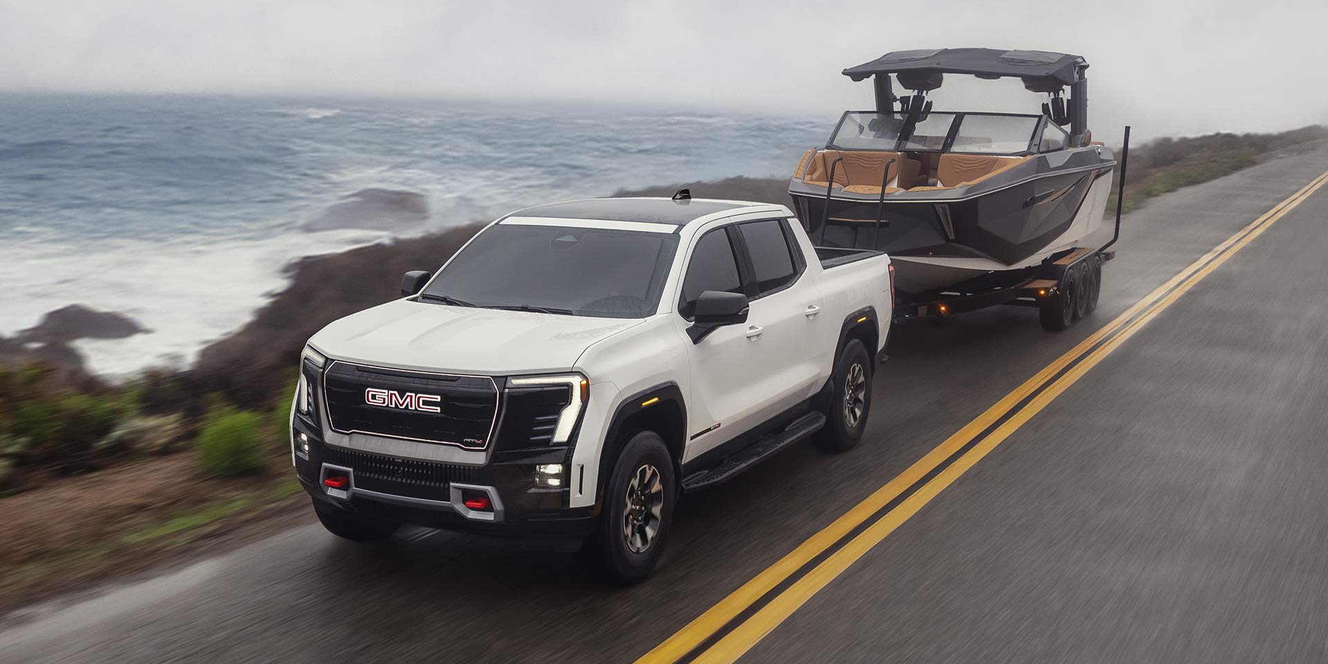 A White GMC Sierra EV Towing a Boat Along a Coastal Highway