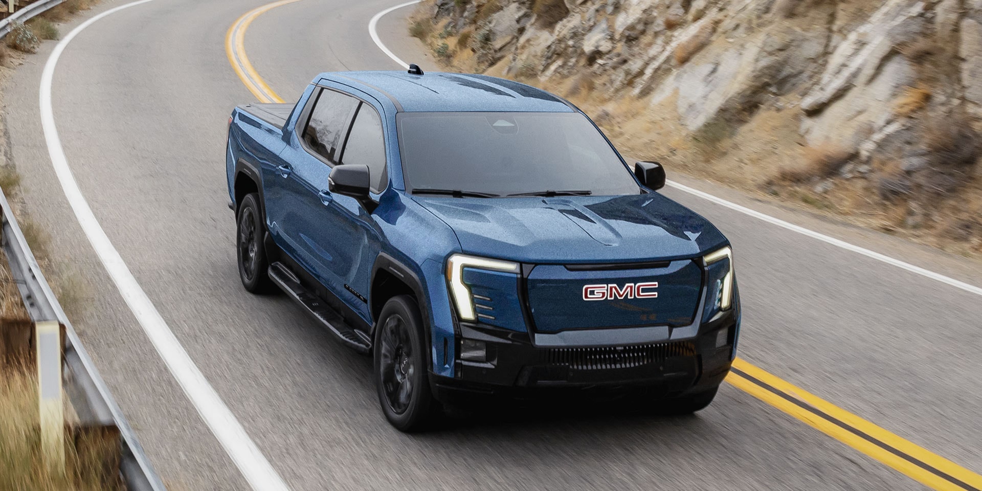A Blue GMC Sierra EV Elevation Cruises on a Winding Road Demonstrating Bold Exterior Styling
