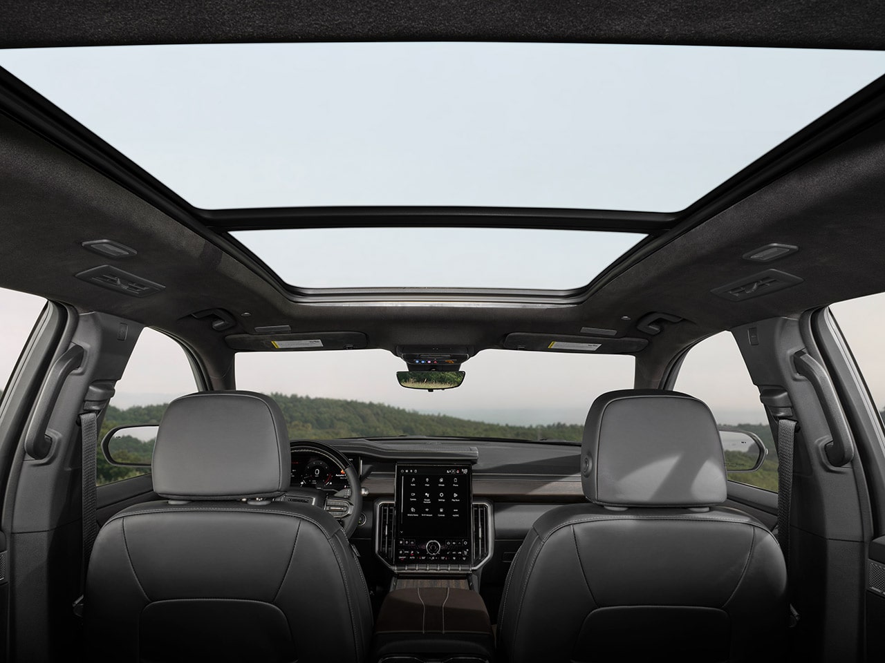 The Interior View of a GMC Vehicle Looking up Through the Panoramic Sunroof, with a Green Landscape Visible in the Distance, along with the Rearview Mirror and Infotainment Screen