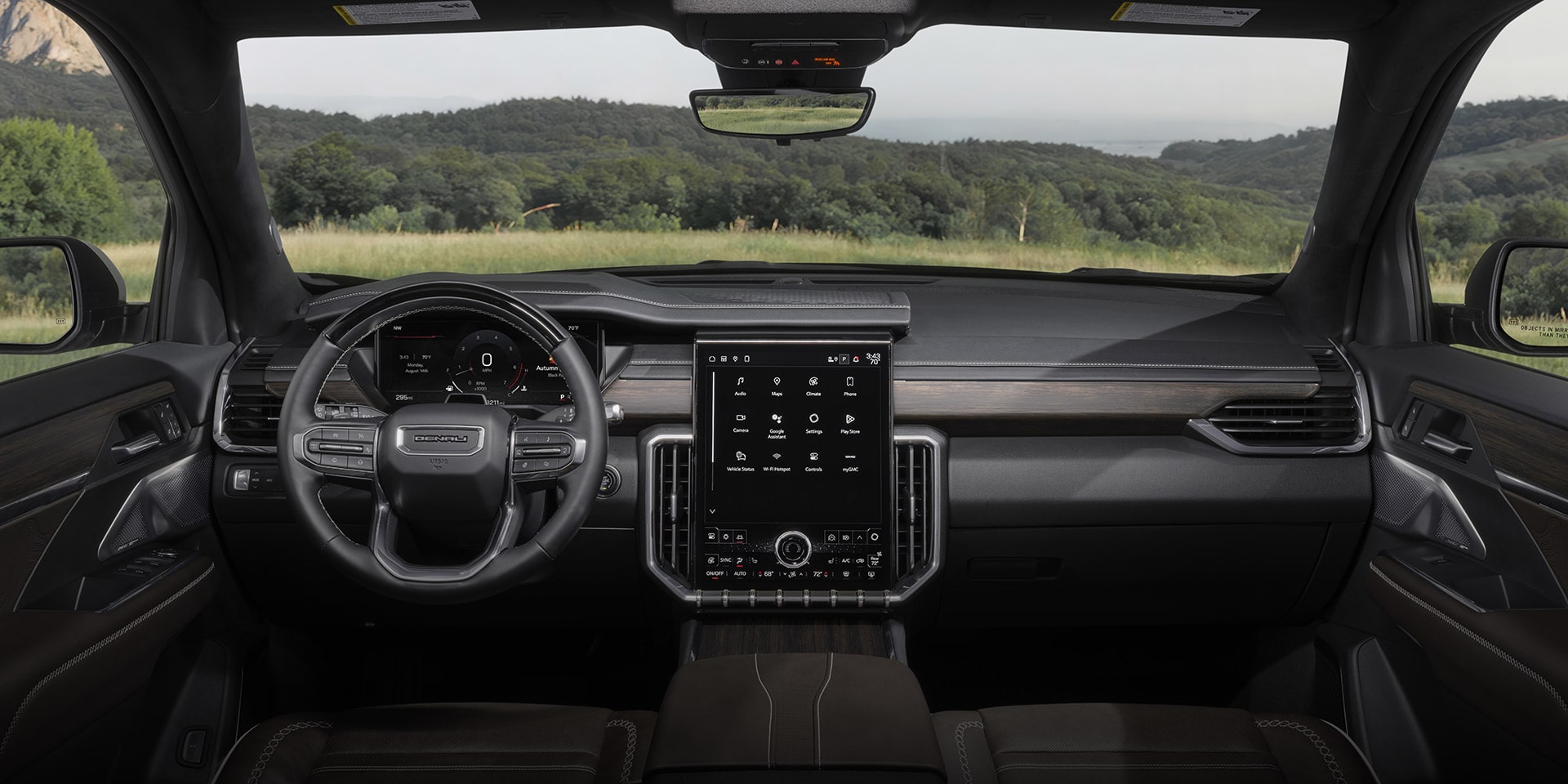 The Interior of a GMC Denali Vehicle Showing the Dashboard, Steering Wheel, and a Large Central Display Screen with App Icons, with a Green Landscape Visible Through the Windshield