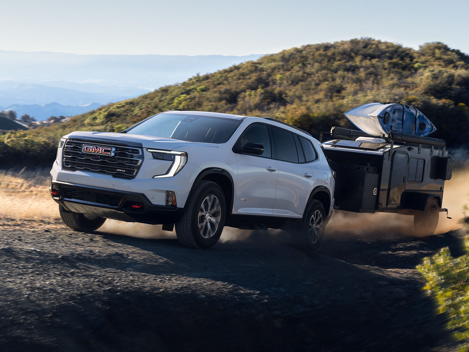 White GMC SUV Pulling a Dark Camping Trailer on a Dusty Dirt Road Through a Hilly Landscape