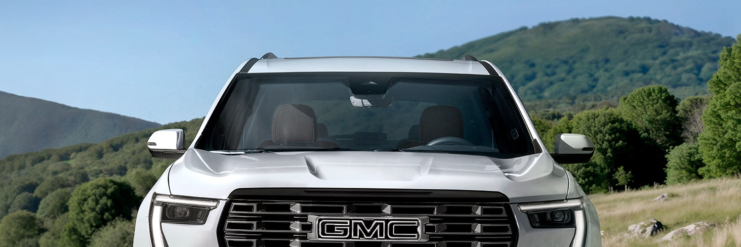Front View of a White GMC SUV With the GMC Logo on the Grille Against a Backdrop of Green Rolling Hills