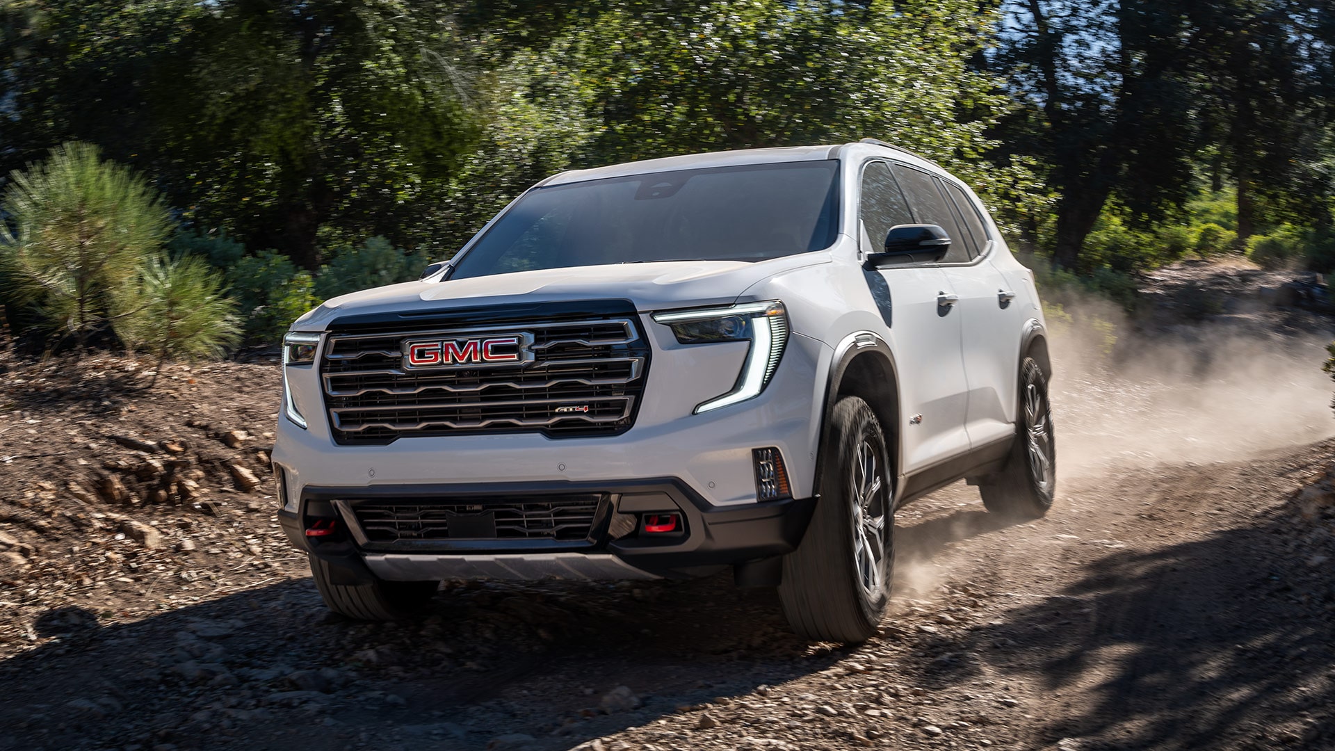 White GMC SUV Driving on a Dusty Dirt Road Through a Forest With Sunlight Filtering Through the Trees