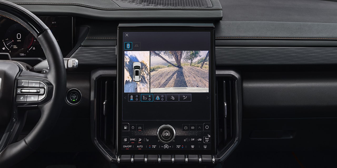 A GMC Vehicle's Infotainment Screen Displaying a Split View of a White SUV From Above and a Side Camera View of a Dirt Road With Tree Shadows
