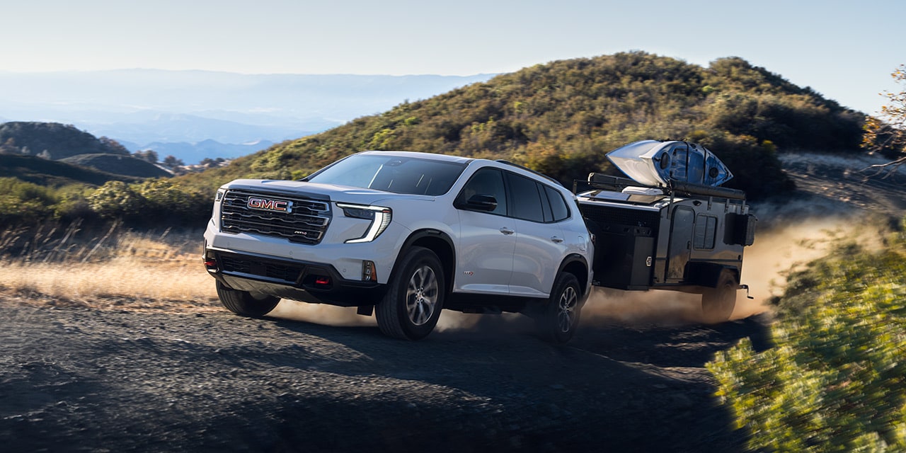 A White GMC SUV Towing a Dark Trailer Up a Dusty, Unpaved Hill With Mountains in The Background Under a Clear Sky