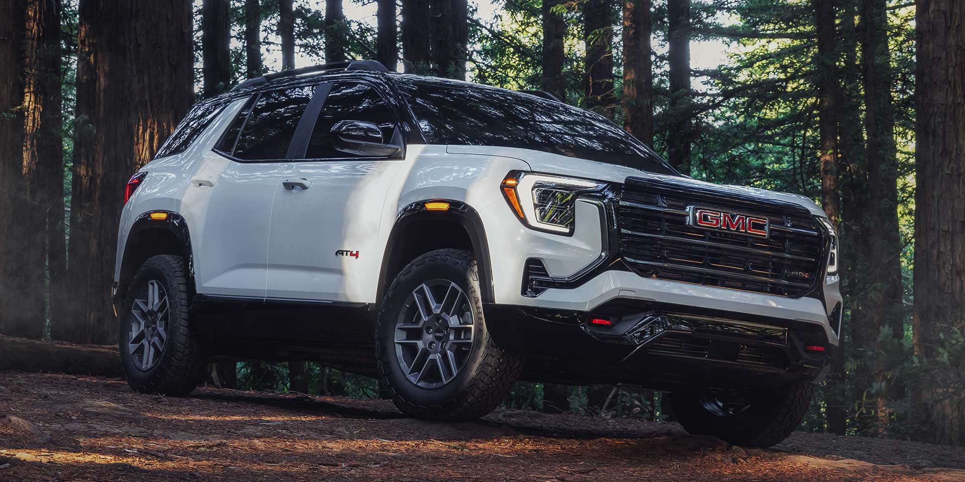 A White GMC AT4 SUV with a Black Roof Parked on a Forest Dirt Path Surrounded by Tall Trees