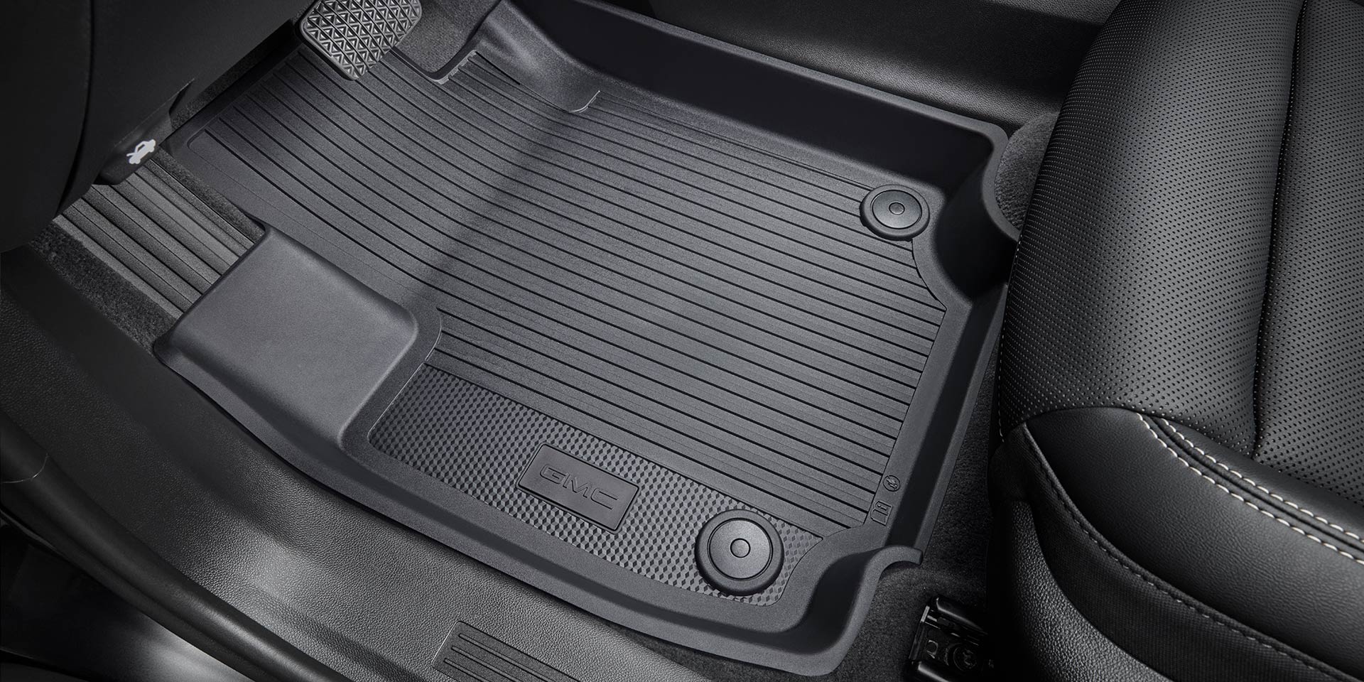 A Close-Up View of a Black GMC All-Weather Floor Mat with a Ribbed Texture and the GMC Logo Embossed on it Next to a Black Leather Seat with White Stitching