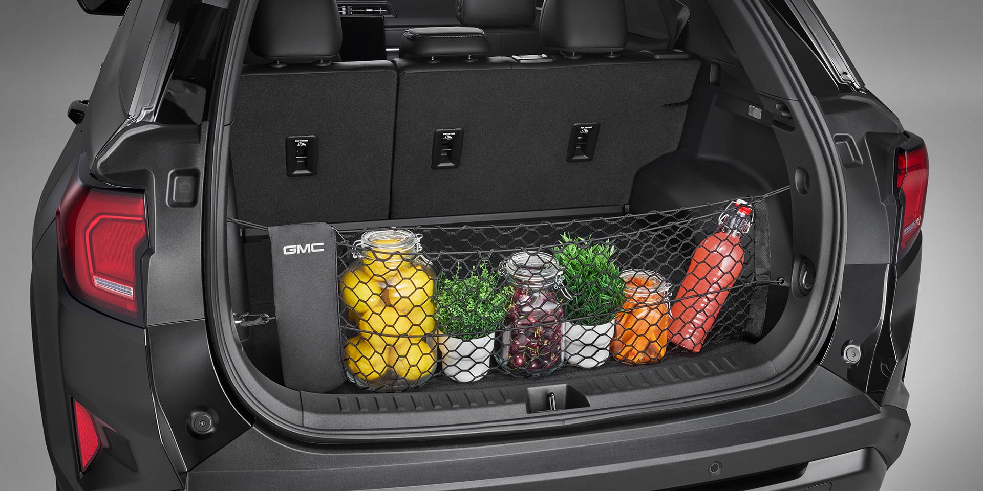 The Open Trunk of a GMC Vehicle Showing a Cargo Net Holding Various Items Like Jars of Lemons, Small Plants, and a Red Water Bottle The GMC Logo is on the Side of the Trunk