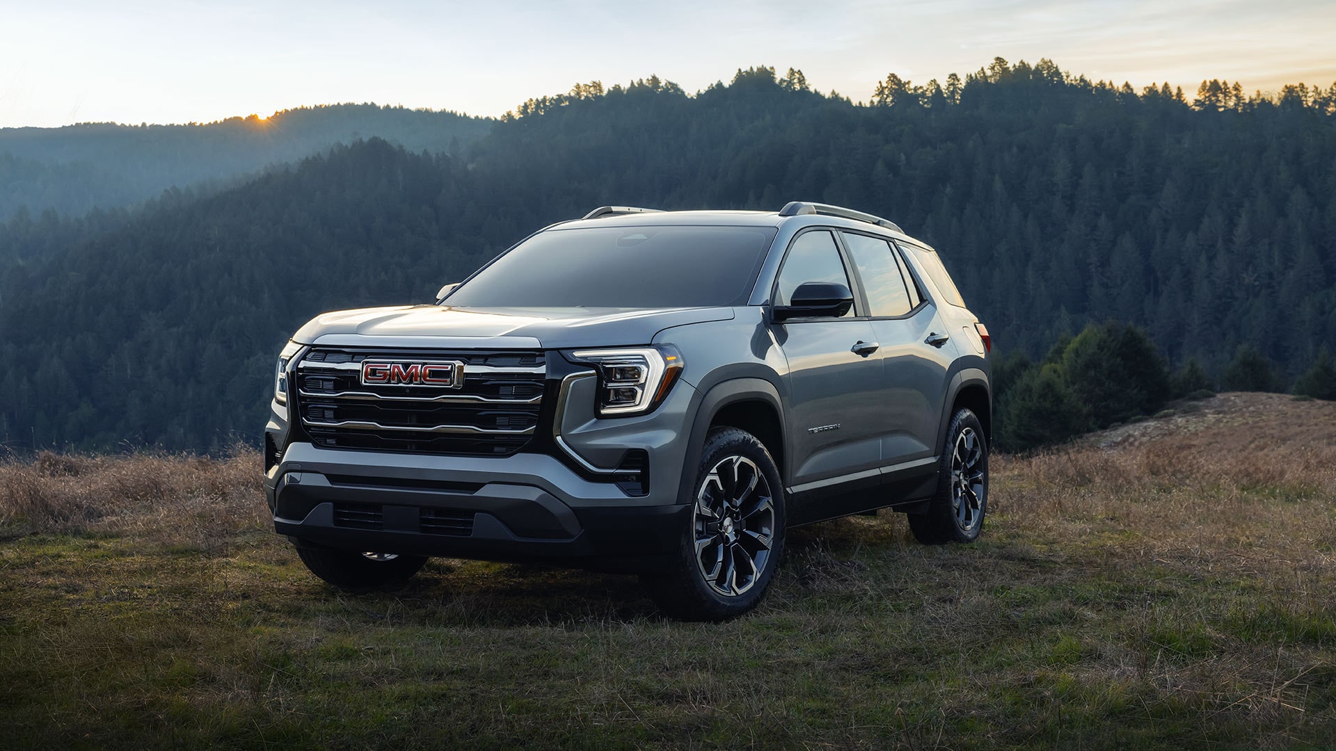 A Gray GMC Acadia AT4 SUV Parked on a Grassy Hill at Sunset With a Forested Mountain Range in the Background