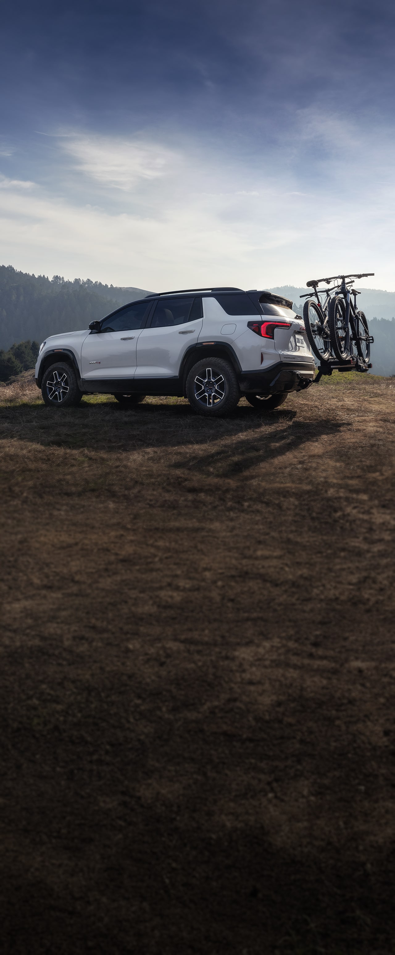 A White GMC Acadia AT4 Parked On a Grassy Hill with Two Mountain Bikes On a Rear Bike Rack
