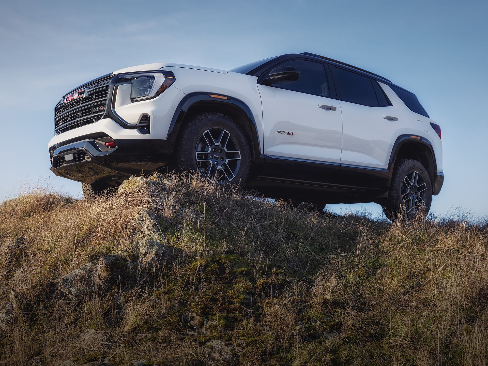 A White GMC AT4 SUV Perched on a Rocky, Grassy Hill against a Clear Blue Sky