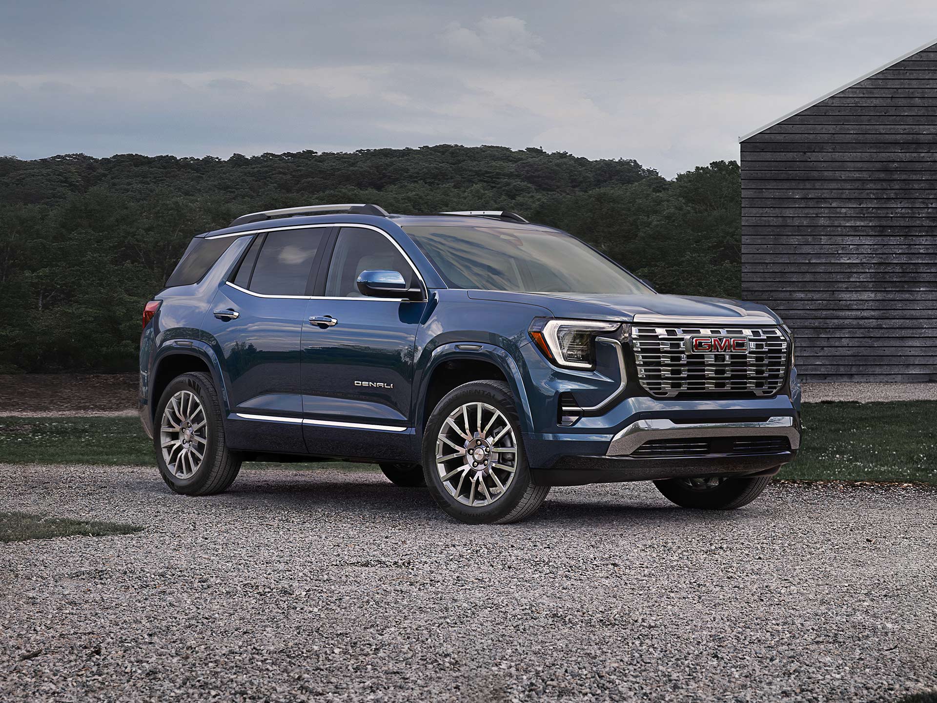 A Blue GMC Denali SUV Parked on a Gravel Lot in Front of a Wooden Building and Trees under a Cloudy Sky