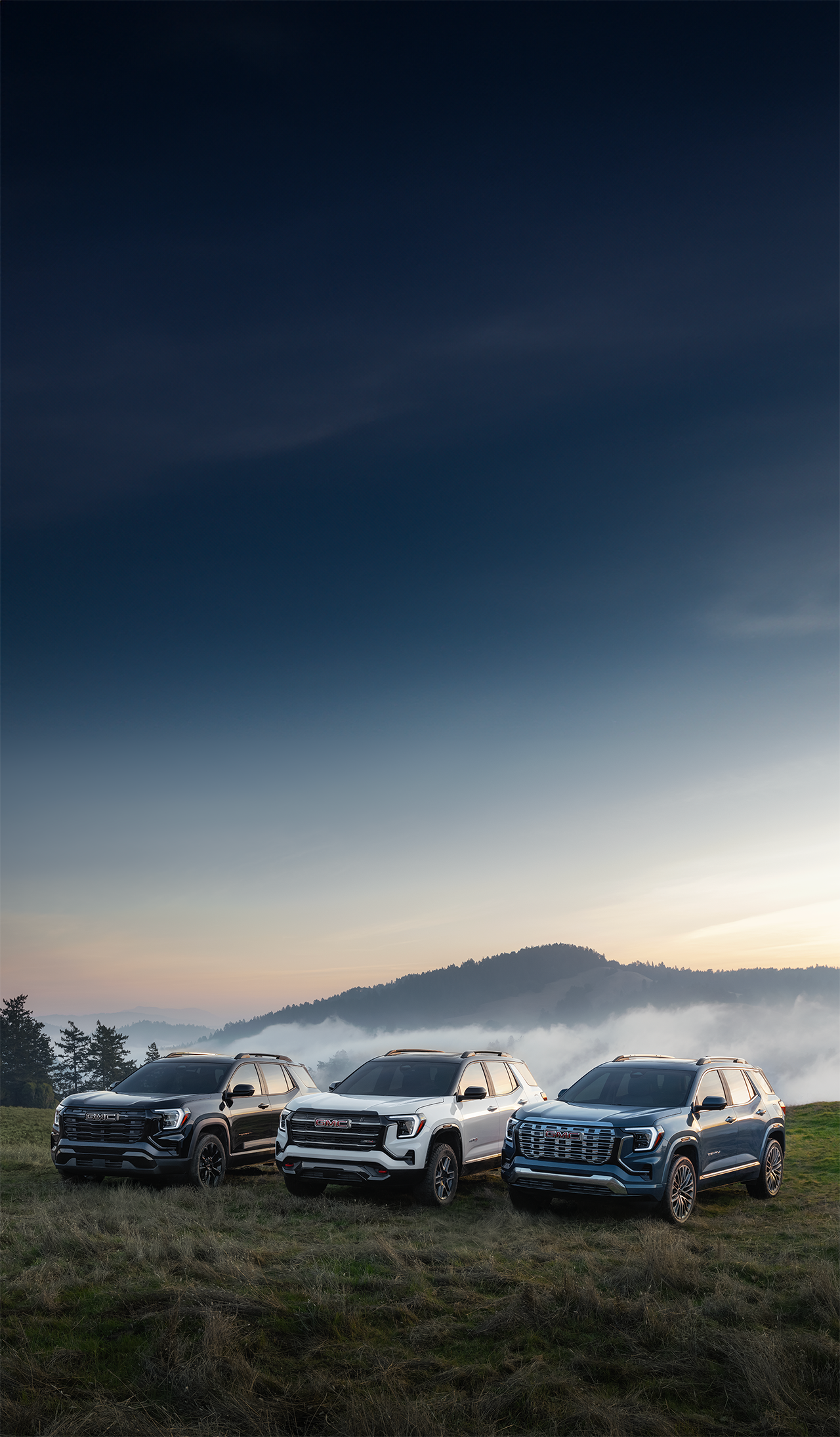 Three GMC Acadia SUVs, a Black, White, and Blue, Parked on a Grassy Hill with Misty Mountains in the Background