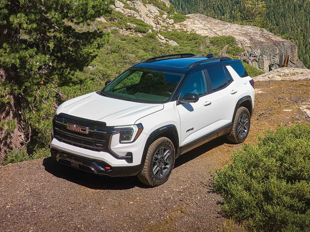 A White GMC AT4 SUV Parked on a Dirt Trail Beside Green Trees And Rocky Terrain Under a Clear Blue Sky