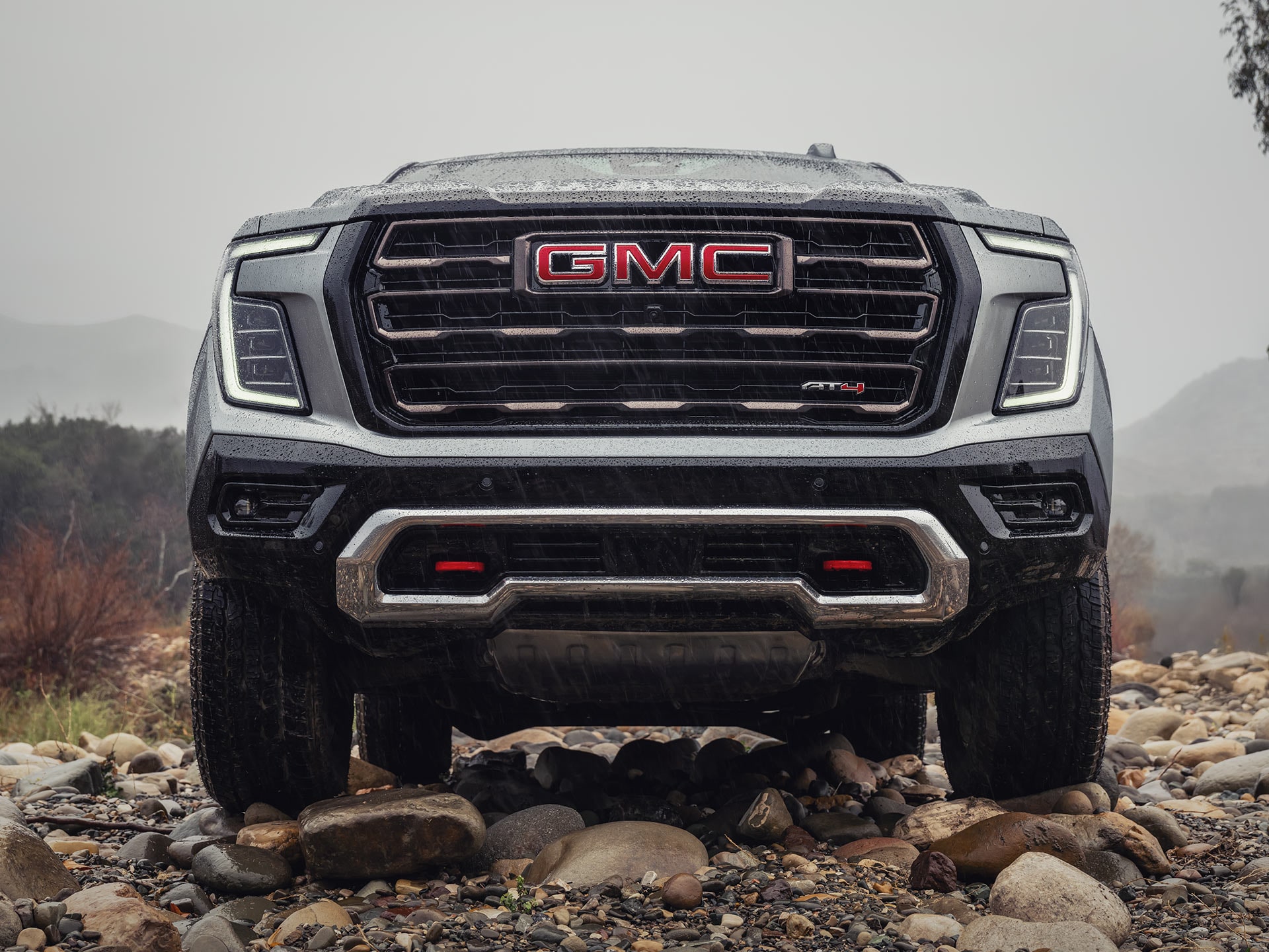 A Close-Up Front View of a Gray GMC Yukon AT4 Grille with Red Recovery Hooks on Rocky Ground