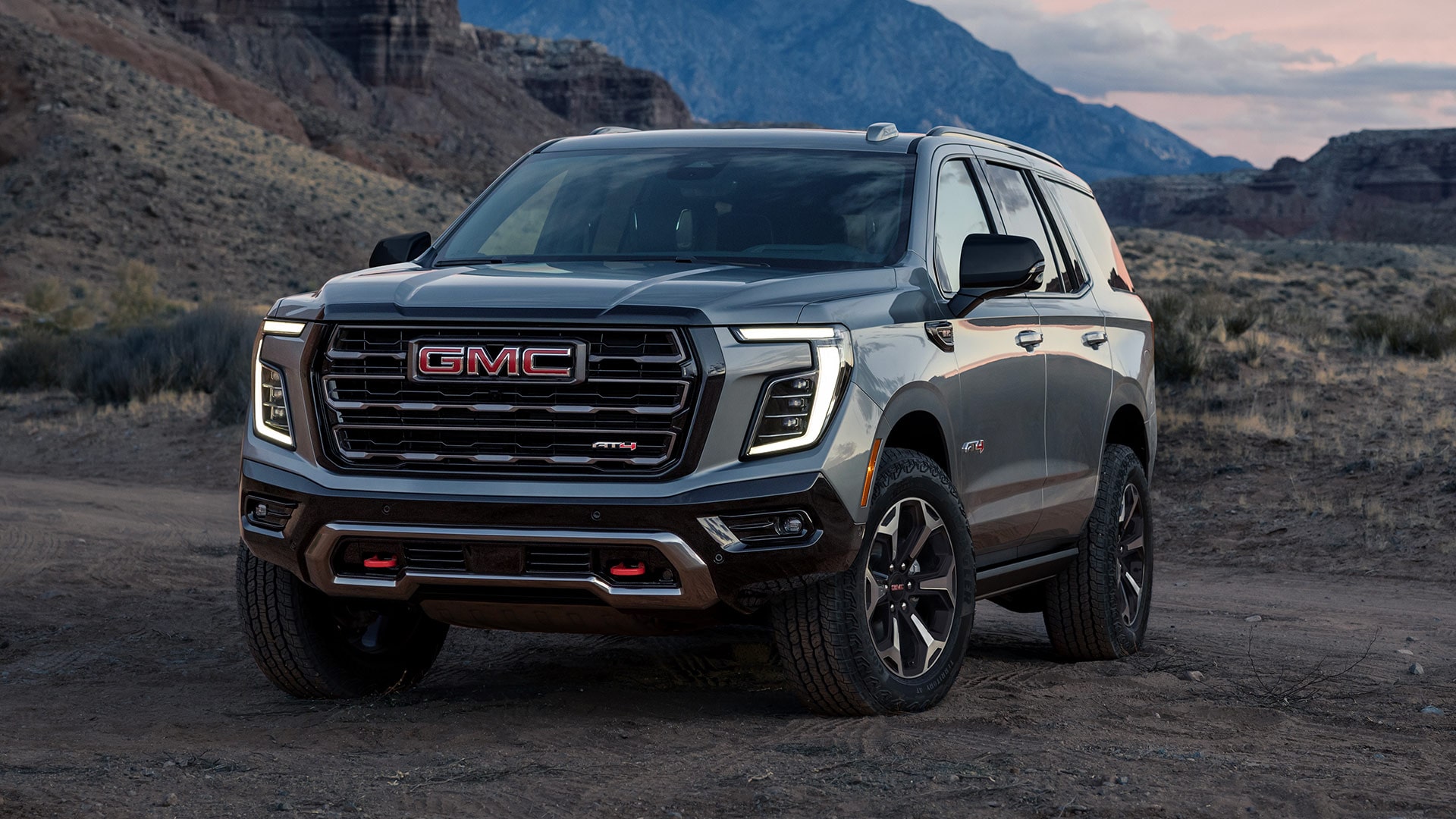 Three-Quarter Front View of a Gray 2027 GMC Yukon AT4 Parked on a Dirt Trail with Rugged Mountains in the Background