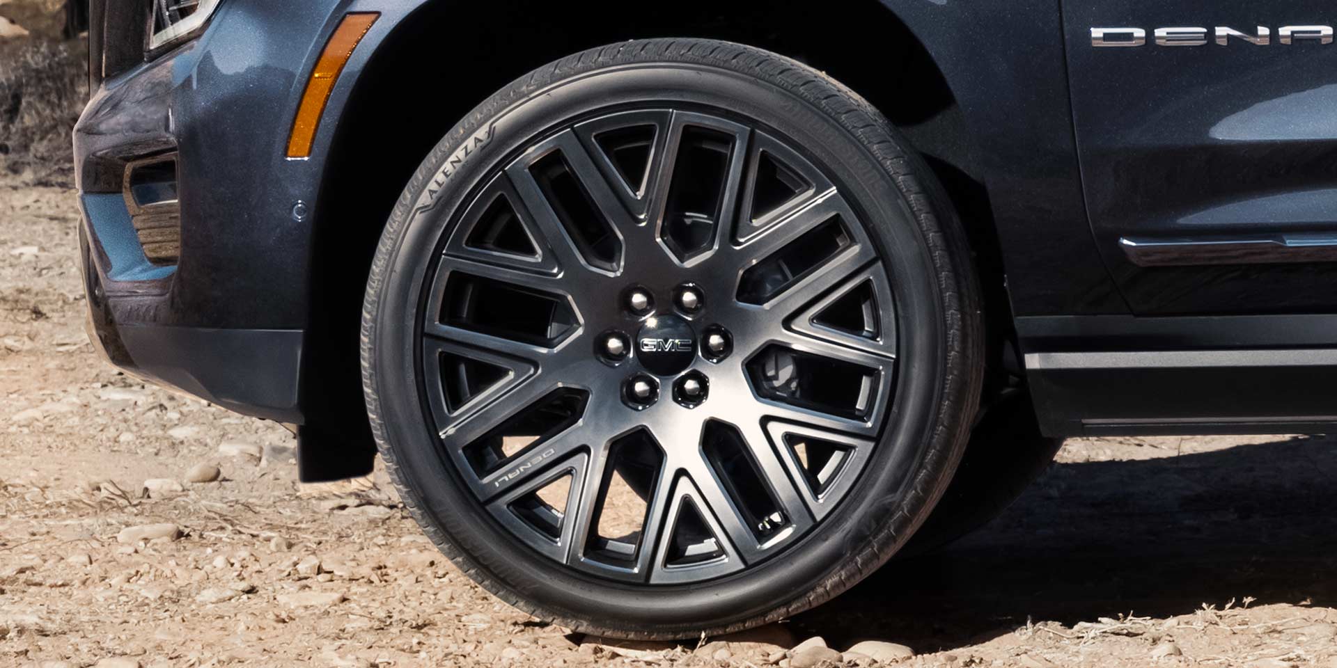 Close-Up of a Dark-Colored 2027 GMC Yukon Denali's Black Multi-Spoke Wheel with Rugged Tire on a Dirt Surface