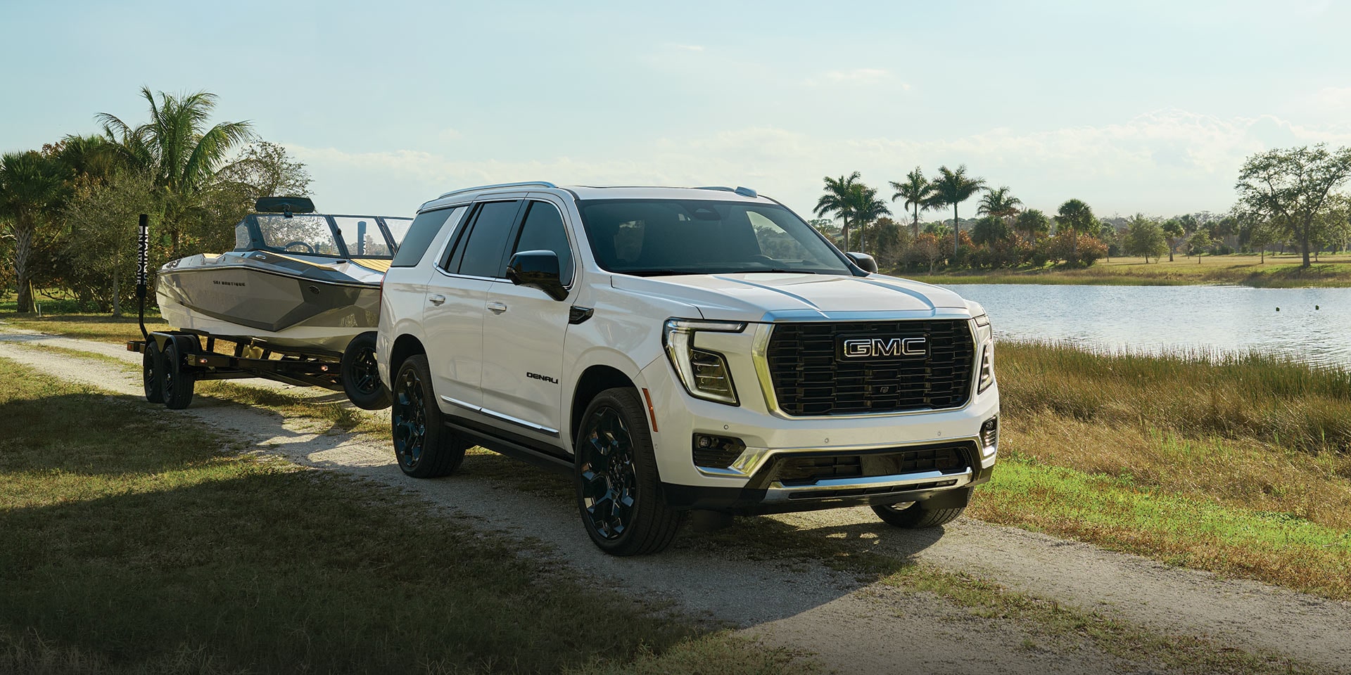 A White 2027 GMC Yukon Denali Pulling a Boat Trailer on a Dirt Road Beside a Tranquil Lake with Palm Trees
