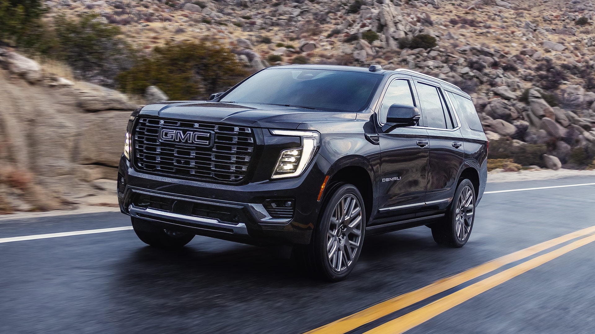 Front Three-Quarter View of a Dark 2027 GMC Yukon Denali Driving on a Winding Road