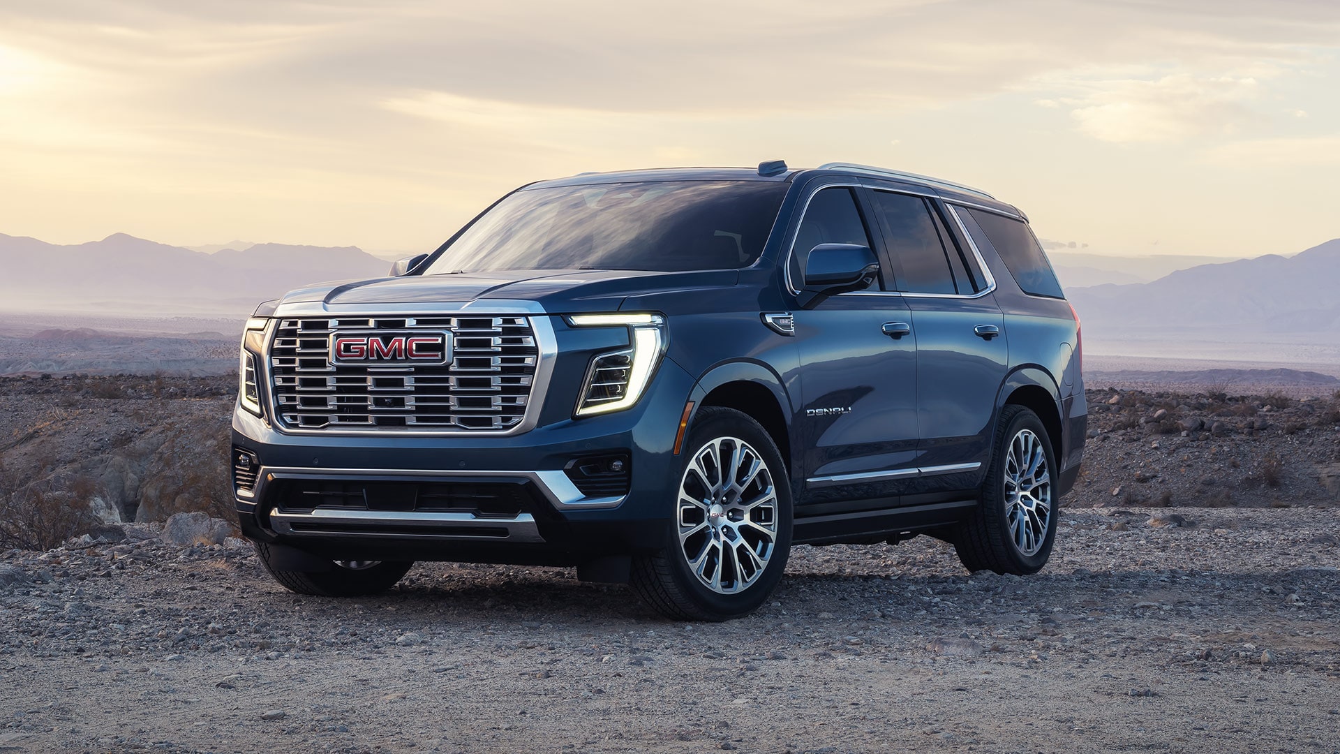 Three-Quarter Front View of a Dark Blue 2027 GMC Yukon Denali Parked on a Dirt Road in a Rocky, Mountainous Landscape