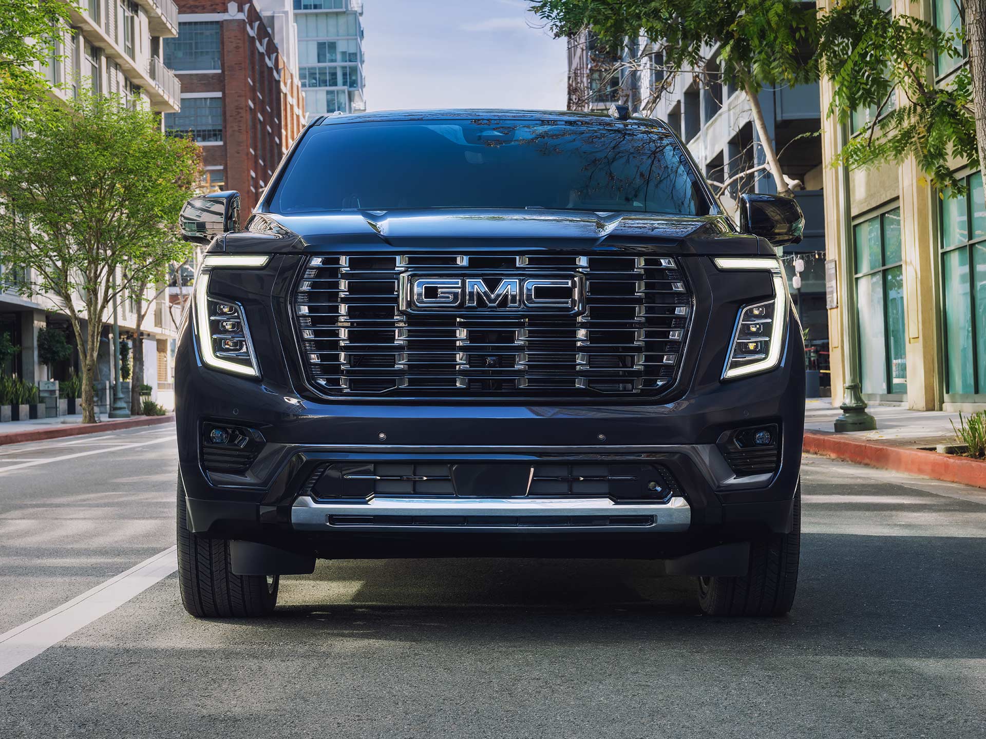Front View of a Dark Blue 2027 GMC Yukon Denali Parked on a Paved City Street with Modern Buildings in the Background