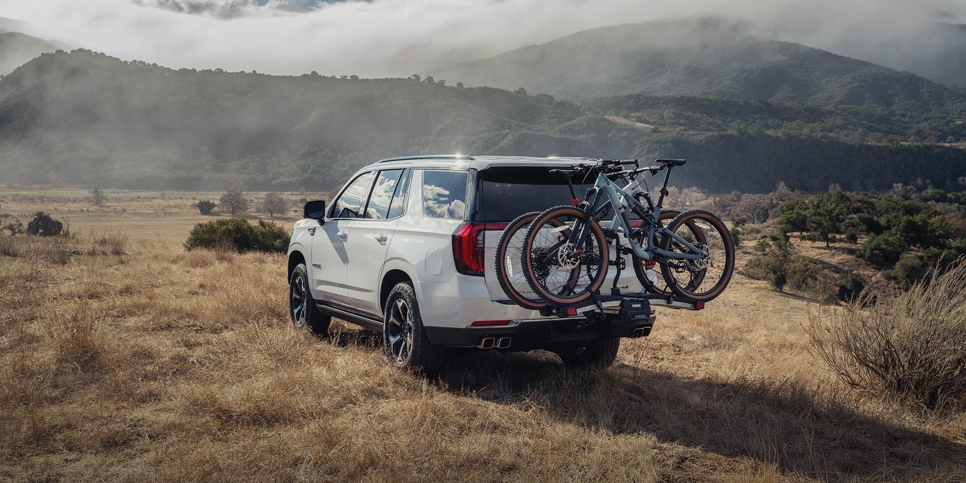 White 2027 GMC Yukon SUV From the Rear with Two Mountain Bikes on a Hitch-Mounted Rack in a Dry Field
