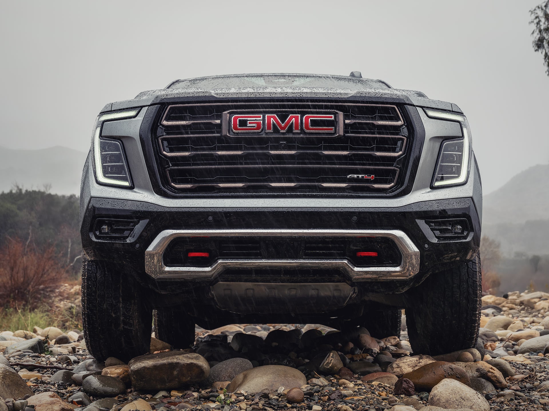 A Close-Up Front View of a Gray GMC Yukon AT4 Grille with Red Recovery Hooks on Rocky Ground