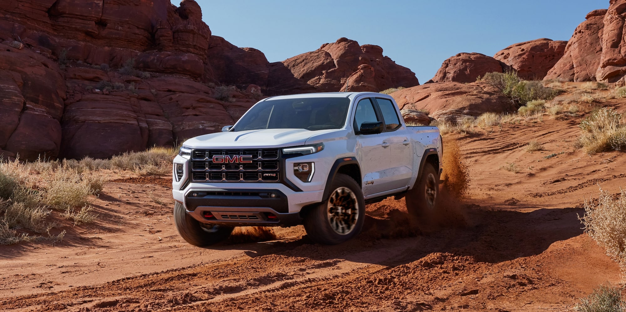 A White 2027 GMC Canyon AT4X Pickup Truck Driving on a Dusty Off-Road Trail in a Desert Landscape