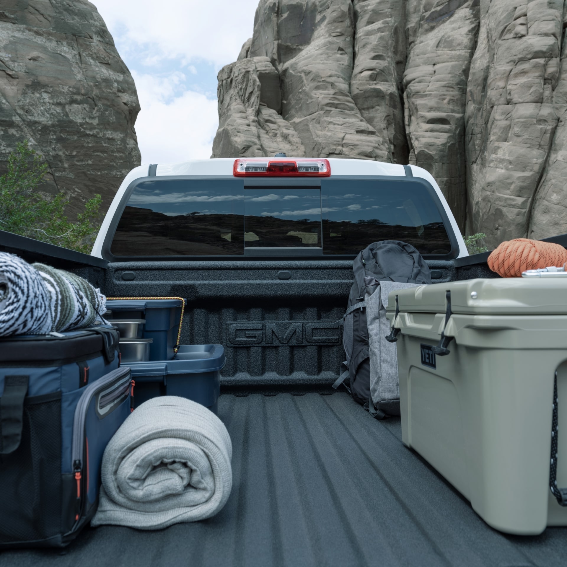 The Bed Of a White 2027 GMC Canyon Truck Filled With Camping Gear Including a Cooler and Rolled Blankets