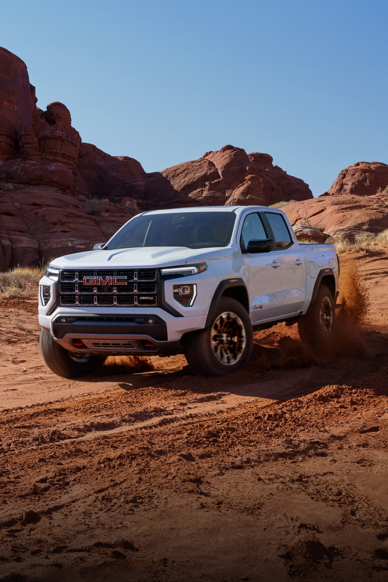 A White 2027 GMC Canyon AT4X Driving on a Dusty Off-Road Trail in a Desert Landscape