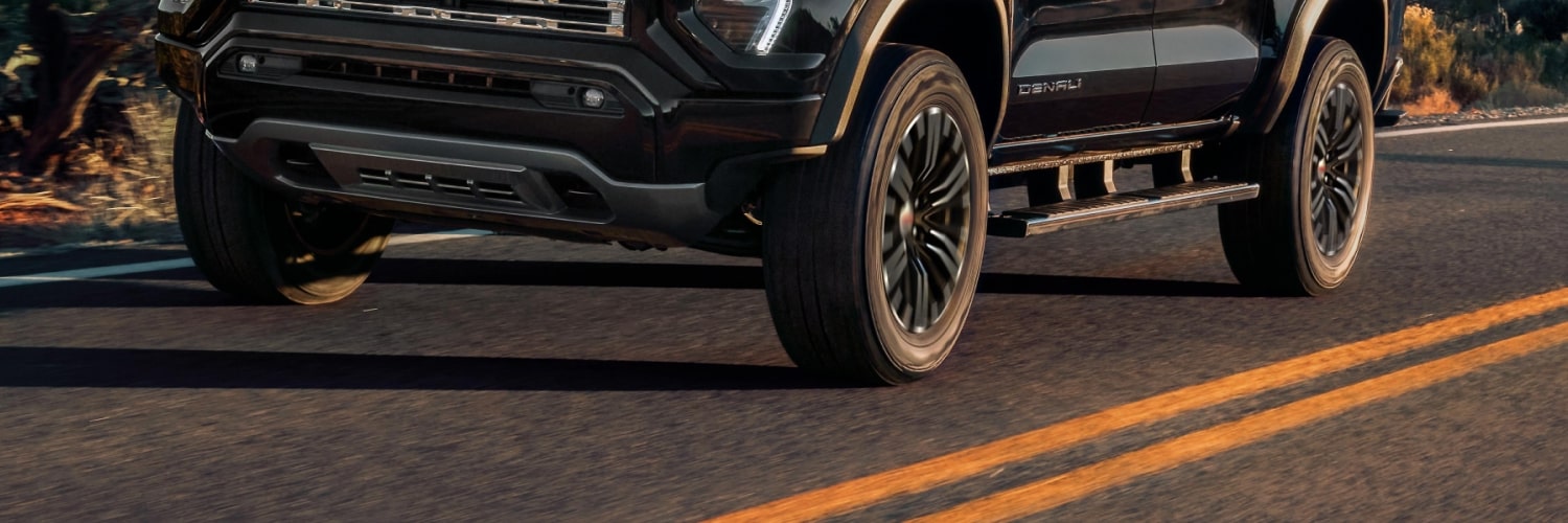 Lower Front and Side View Of a Black 2027 GMC Canyon Denali Truck With Running Boards and Dark Wheels on a Road
