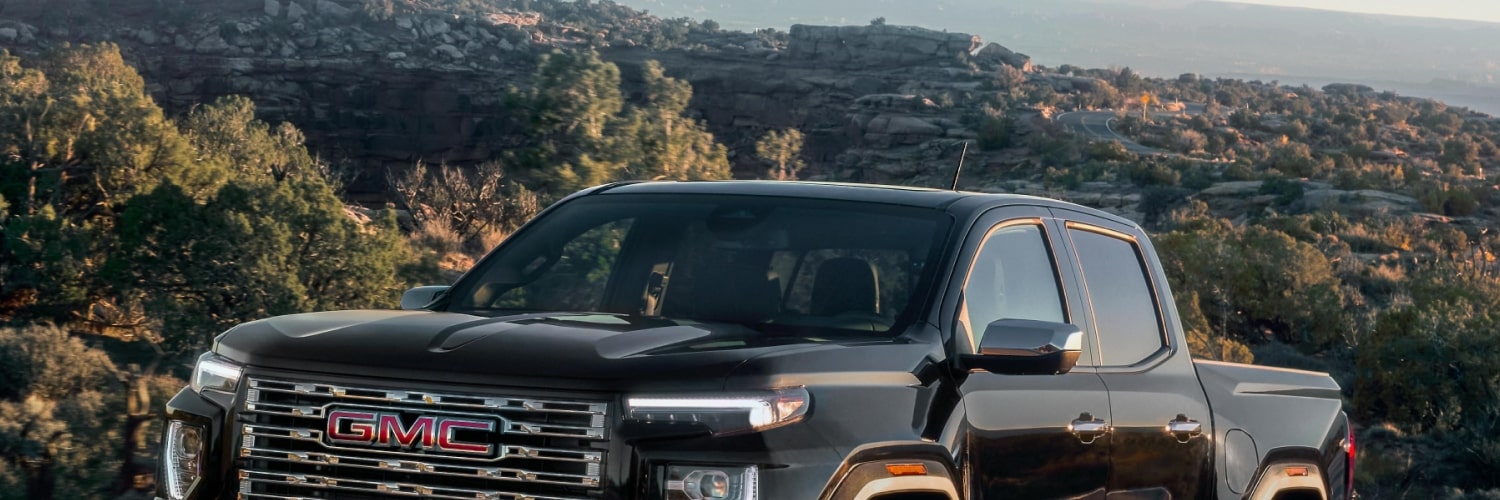Upper Front View Of a Black 2027 GMC Canyon Truck Driving on a Winding Road With Mountainous Terrain in the Background