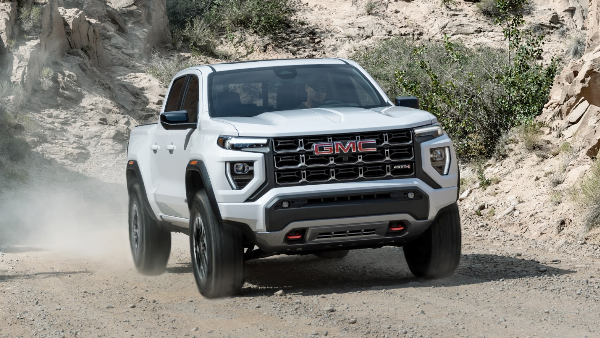 A White 2027 GMC Canyon AT4X Truck Driving on a Dusty Off-Road Trail Surrounded by Rocks