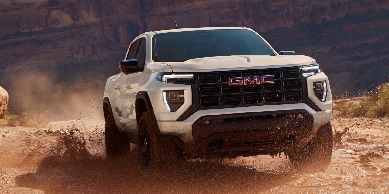 A White 2027 GMC Canyon AT4X with the GMC Logo on the Grille Driving on a Dusty Off-Road Trail
